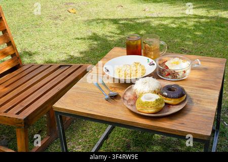 Verschiedene süße und herzhafte Gerichte, darunter Donuts, cremige Pasta, eine Reisschale und Eistee, die auf einem Außentisch des Cafés serviert werden. Stockfoto