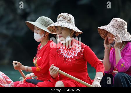 Drei Frauen mit Hüten und Masken segeln auf einem Boot. Sie sind in traditioneller roter und rosa Kleidung gekleidet und genießen einen sonnigen Tag. Trang An Stockfoto