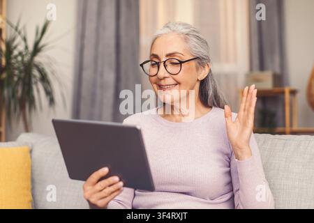 Zufriedene ältere Frau mit grauen Haaren, die zu Hause ein Tablet und Videotelefonie verwendet, Gesundheitskonzept Stockfoto
