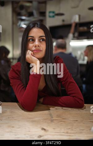 Junge Frau mit langen dunklen Haaren, die nachdenkt und ihr Kinn auf eine Hand stützt, an einem Holztisch Stockfoto