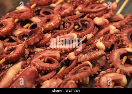 Eine Nahaufnahme von saftigen Oktopus-Tentakeln, die auf Holzspieße gefädelt und über heißer Holzkohle gegrillt werden. Das rötlich-braune Meeresfrüchtegericht glänzt mit Öl, sh Stockfoto