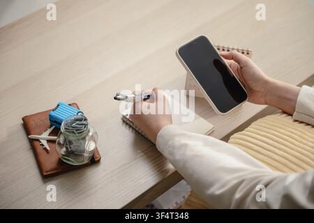 Frau hält Mobiltelefon mit leerem Bildschirm, Schreibnotizbuch, Reisebudgetplanung in der Nähe eines Geldspardose-Schreibtischkalenders. Konzept der Urlaubsfinanzierung Stockfoto