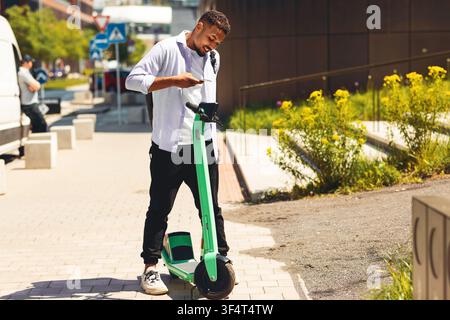 Junger schwarzer Mann, der ein Smartphone benutzt, während er auf einem Elektroscooter in einer städtischen Umgebung steht Stockfoto