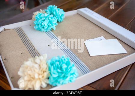 Schräge Nahaufnahme eines rustikalen mit Sackleinen ausgekleideten Geschenktabletts, dekoriert mit türkisfarbenen Papierblumen, gestreiften Bandakzenten und einem leeren weißen Notizzettel Stockfoto