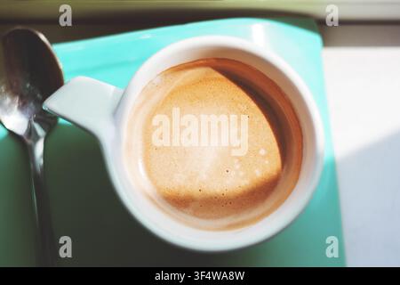 Ein erfrischender heißer Espresso, gekrönt mit reichem Schaum, serviert in einer kleinen weißen Keramiktasse neben einem Metalllöffel, platziert auf einem türkisfarbenen Tablett. Stockfoto