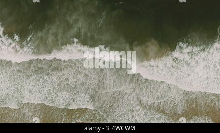 Luftaufnahme von Meereswellen, die auf die sandige Küste schlagen und brechen, mit weißem Seeschaum und Mustern im Wasser Stockfoto