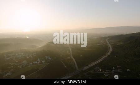 Luftbild einer sich durch eine Tallandschaft windenden Autobahn mit Nebel und Morgensonne über Feldern und einer kleinen Stadt Stockfoto