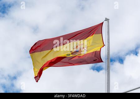 Die Nationalflagge Spaniens mit ihren roten und gelben Streifen und dem Wappen weht an einer Flaggenstange vor einem blauen Himmel mit weißen Wolken Stockfoto