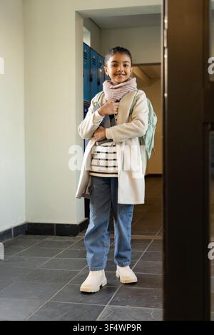 Afroamerikanische Schülerin steht im Flur und hält einen mintgrünen Rucksack, blaue Spinde im Hintergrund Stockfoto
