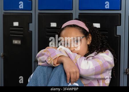 Afroamerikanisches Vorschulmädchen sitzt auf dem Schulboden neben den Spinden, umarmt ihre Knie und trägt eine Brille Stockfoto
