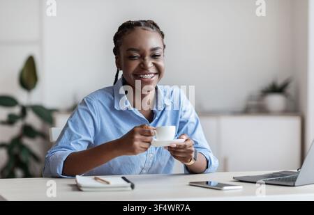 Fröhliche Afroamerikanische Frau Genießen Kaffee Am Arbeitsplatz In Weiß Büro Stockfoto
