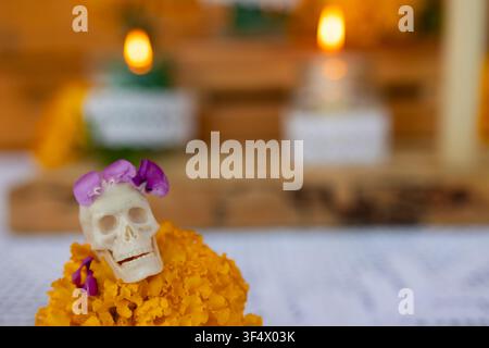 Nahaufnahme eines kleinen Zuckerschädels, der mit violetten Blütenblättern verziert ist und auf einer leuchtenden Ringelblumenblüte auf dem Altar des Tages der Toten ruht, mit warmen verschwommenen Kerzen, rustikalem Holz und weichem Bokeh im Hintergrund Stockfoto