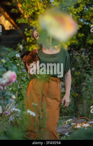 Mädchen-Bauernhofbesitzerin hält braune Henne im Hinterhofgarten fest. Fröhliche Bäuerin hält Henne in ihren Händen auf dem Bauernhof. Stockfoto