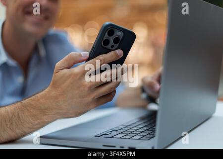 lächelnder Mann, der ein Smartphone benutzt, während er an seinem Laptop arbeitet. Nahaufnahme von Händen, die das Telefon auf einem weißen Schreibtisch halten, mit unscharfem warmem Bokeh-Hintergrund. Pe Stockfoto