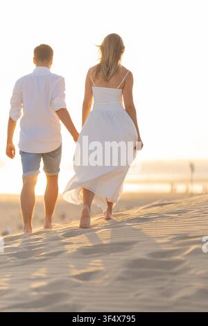 Junges Paar geht Hand in Hand auf dem Strandsand bei Sonnenuntergang in der Nähe der Meereswellen Stockfoto