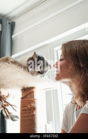 Frau lächelt ihre graue und weiße Katze an, die auf einem Katzenbaum in der Nähe eines Fensters entspannt. Warme Interaktion fängt Zuneigung, Kameradschaft und Alltagsleben ein Stockfoto