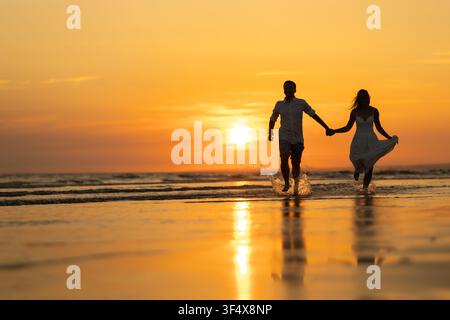 Entzückendes Paar genießt einen Sonnenuntergangsspaziergang am Strand, während es Händchen hält und im Wasser planscht Stockfoto
