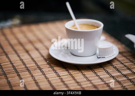 Eine Nahaufnahme einer kleinen weißen Keramiktasse, die mit heißem Espresso auf einer Untertasse gefüllt ist, begleitet von einem Plastikrührer, einem Zuckerpäckchen und einer Milchkapsel, Ort Stockfoto