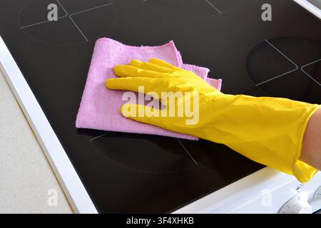 Person in gelbem Handschuh reinigt eine schwarze Keramik-Kochplatte mit einem rosa Tuch Stockfoto