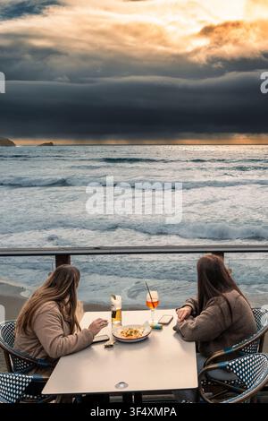 Zwei Freundinnen sitzen auf der Außenterrasse der Fistral Boardmasters Beach Bar, essen und trinken Alkohol und beobachten nahenden Regen Stockfoto