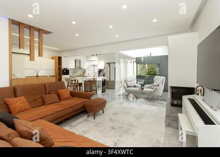 Geräumiges modernes Wohnrauminterieur im offenen Grundriss mit einer großen Ledersofaecke, weißem Klavier, Kamin, Essbereich und offener Küche Stockfoto