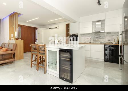Modernes Wohnkücheninterieur mit eleganten weißen Schränken, Marmorfliesenboden und einer zentralen Kücheninsel mit einem Weinkühlschrank Stockfoto