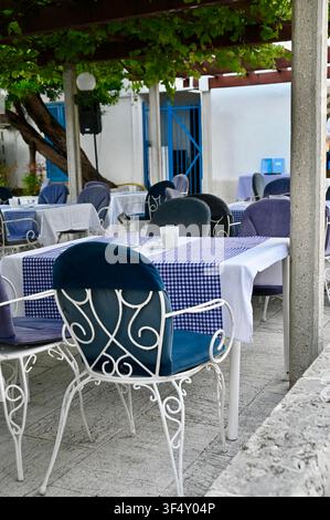 Ein Blick auf die Außenterrasse einer traditionellen griechischen Taverne. Die Szene zeigt weiße schmiedeeiserne Stühle mit blauem Samtpolster, die um einen Tisch mit einer blau-weißen Musterdecke angeordnet sind. Die Tische stehen unter einer mit üppigen Weinreben bedeckten Holzpergola auf einem steinernen Pflasterboden. Im Hintergrund ist die charakteristische weiß-blaue griechische Architektur zu sehen. Stockfoto