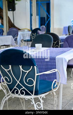 Ein Blick auf die Außenterrasse einer traditionellen griechischen Taverne. Die Szene zeigt weiße schmiedeeiserne Stühle mit blauem Samtpolster, die um einen Tisch mit einer blau-weißen Musterdecke angeordnet sind. Die Tische stehen unter einer mit üppigen Weinreben bedeckten Holzpergola auf einem steinernen Pflasterboden. Im Hintergrund ist die charakteristische weiß-blaue griechische Architektur zu sehen. Stockfoto