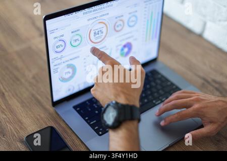 Nahaufnahme eines Geschäftsmannes, der auf ein Finanzanalysen-Dashboard auf einem Laptop-Bildschirm zeigt. Holzschreibtisch-Arbeitsplatz mit einem Smartphone und einem weißen Ziegelwa Stockfoto