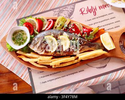 gegrillter Fisch mit Beilagen auf einem Holzteller, im Hintergrund eine Fischkarte "Riblji Menu" in südslawischer Sprache Stockfoto
