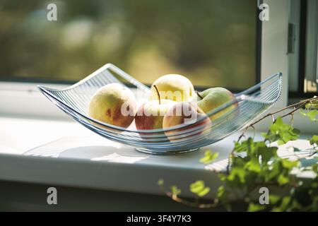 Frische reife Äpfel arrangiert in einer modernen Glas-Fruchtschale auf einer weißen Fensterbank mit natürlichem Licht und grünen Blättern im Vordergrund, die eine gesunde Stockfoto