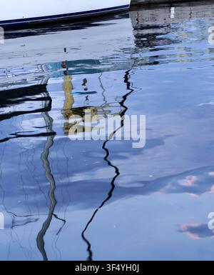 Bootsmasten und Rümpfe erzeugen verzerrte, farbenfrohe Spiegelungen auf der ruhigen, sanft wogenden Wasseroberfläche in einem Hafen, was eine abstrakte und friedliche Atmosphäre schafft Stockfoto