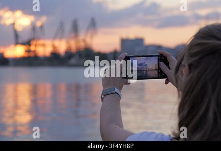 Frau hält ein Smartphone und dokumentiert das schöne Sonnenlicht, das sich auf der Wasseroberfläche eines Hafens mit industriellen Kränen im Hintergrund spiegelt Stockfoto