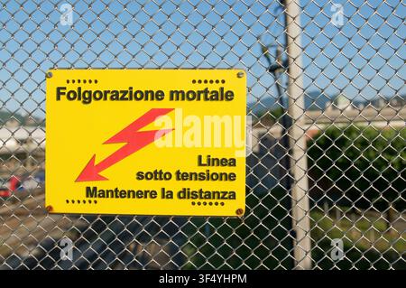 Nahaufnahme eines gelben Hochspannungs-Warnschilds mit rotem Blitzsymbol, das lautet Folgorazione mortale (tödlicher Stromschlag) und an einem Zaun montiert ist Stockfoto