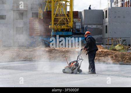 Ein Arbeiter mit einem orangefarbenen Helm schneidet Beton mit einer staubigen Schleifmaschine auf einer Baustelle mit einem gelben Turmdrehkran und einem unfertigen Wohnblock Stockfoto