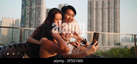 Verschiedene Freunde lächeln, machen eine Herzensgeste, verwenden ein Smartphone für ein Selfie auf dem Stadtdach Stockfoto
