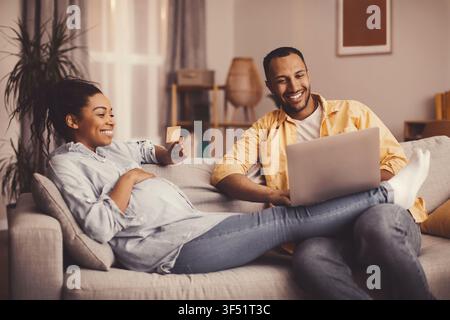 Zufriedene afroamerikanische schwangere Frau, die online mit ihrem Partner zu Hause einkauft, Familienkonzept Stockfoto