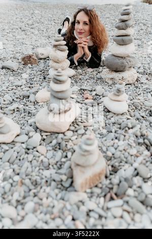 Kiesstrand-Steine: Lächelnde Frau liegt an einem felsigen Ufer, umgeben von balancierten Steinpyramiden, und genießt einen friedlichen Moment der Achtsamkeit Stockfoto