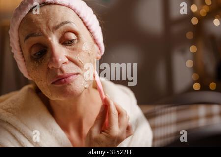 Reifes Hautpflege-Konzept. Ältere Seniorin mit Stirnband macht eine Gesichtsmasage mit organischem Gesichtsscrub, trägt Scrub-Creme auf, peelt ihre Gesichtshaut Stockfoto