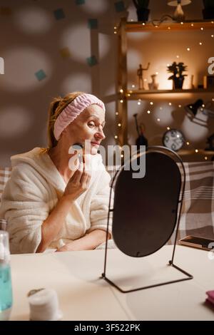 Reife Frau in Bademantel und Stirnband verwendet einen Jade-Roller während einer Gesichtsmasage und schaut in den Spiegel im häuslichen Innenraum Stockfoto