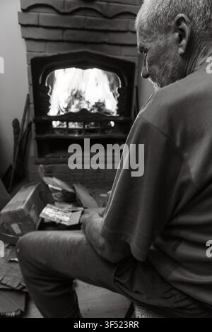 Alter Mann sitzt allein vor dem Kamin, Schwarz-Weiß. Seniorenleben. Alter müder Mann beobachtet die Flamme im Kamin, Monochrom. Stockfoto