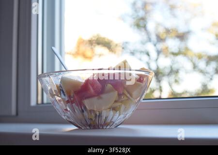 Ein frischer gemischter Obstsalat, serviert in einer dekorativen Glasschale, die auf einer Fensterbank platziert ist, mit Stücken von Wassermelone und Birnen, die in natürlichem Licht baden Stockfoto
