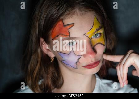 Nahaufnahmeporträt eines Mädchens im Vorpubertätsalter mit bunten Sternen im Gesicht, das gegen einen dunklen Hintergrund aufblickt Stockfoto