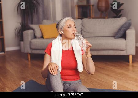 Aktive Seniorin kaukasischer Abstammung trinkt Wasser nach dem Heimtraining, Gesundheitskonzept Stockfoto