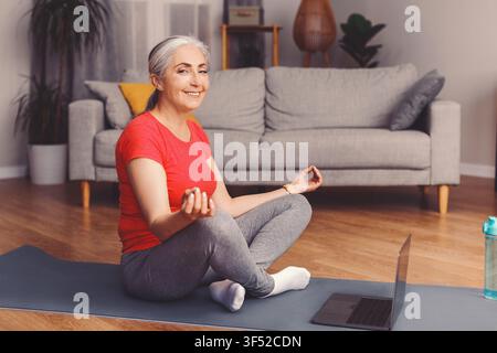 Lächelnde ältere Frau, die zu Hause Yoga praktiziert, glücklicher und entspannter Zustand, Wellness-Konzept Stockfoto