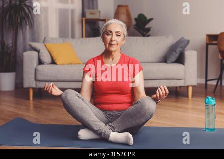 Seniorin praktiziert Meditation im Wohnzimmer, konzentriertes und ruhiges mentales Wohlbefinden Konzept Stockfoto