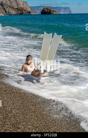 Spielmoment im Ozeanwellenbrecher festgehalten, während ein lächelnder Schwimmer mit langen Flossen an einer kiesigen Strandküste schwimmt Stockfoto