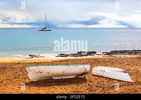 Zwei verwitterte Dingis ruhen auf einem sandigen Strand, während ein Katamaran vor der Küste vor Anker liegt, unter einem düsteren bewölkten Himmel über ruhigem türkisfarbenem Wasser Stockfoto