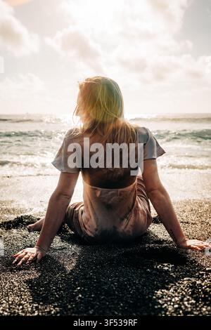 Frau in einem Kleid sitzt auf dunklem Sandstrand und betrachtet den Ozean Stockfoto