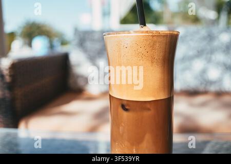 Eine Nahaufnahme eines kalten Eiskaffees in einem hohen Glas mit einer dicken, schaumigen Schaumschicht oben und einem eingesteckten Strohhalm, der auf einem Tisch in G platziert ist Stockfoto